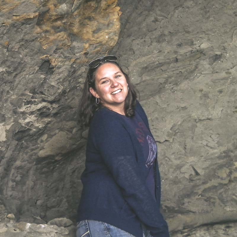 Portrait of a smiling woman with sunglasses perched on her head, posing in front of a rugged rock face, wearing a casual navy blue sweater, exuding a joyful and relaxed outdoor vibe.