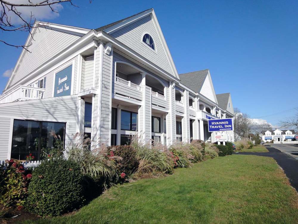 Exterior view of the Hyannis Travel Inn on a sunny day, featuring a white multi-story building with blue signage, a covered porch, and well-maintained landscaping along a quiet street.
