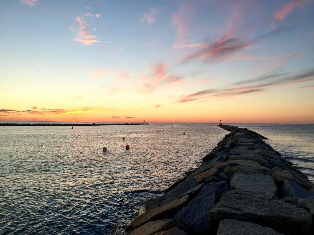 This image captures a serene sunset over Cold Storage Beach, with a rocky jetty extending into calm waters under a sky painted with soft hues of pink, orange, and blue. The tranquil ocean reflects the warm colors, while scattered buoys add subtle details to the scene. It's a peaceful and picturesque coastal moment, perfect for evening relaxation.