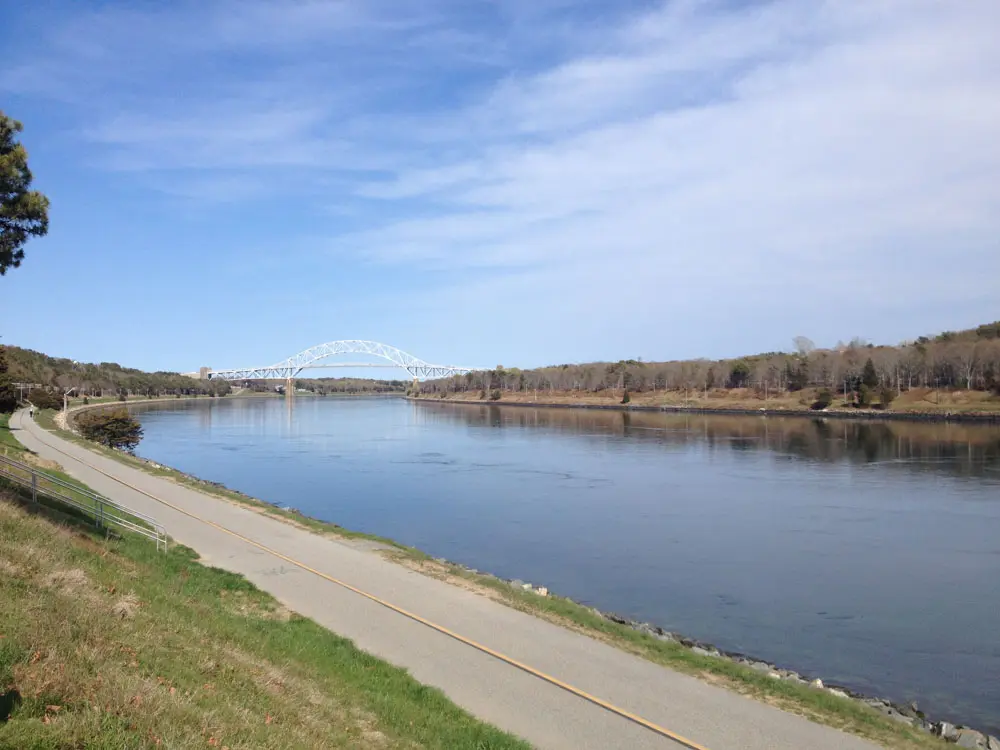 This image showcases a serene view of the Cape Cod Canal with a paved pathway running alongside the calm water, perfect for walking or biking. In the distance, a large arched bridge spans the canal, surrounded by sparse woodland and under a partly cloudy blue sky. The peaceful scene highlights the charm of this historic and scenic area.