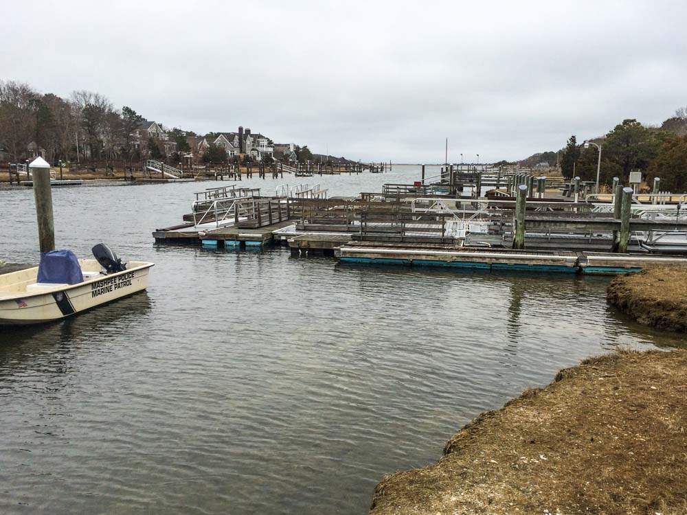 Overcast day at a marina with a Mashpee Police Marine Patrol boat moored beside several empty boat docks, reflecting a quiet off-season atmosphere in a waterway surrounded by residential houses.