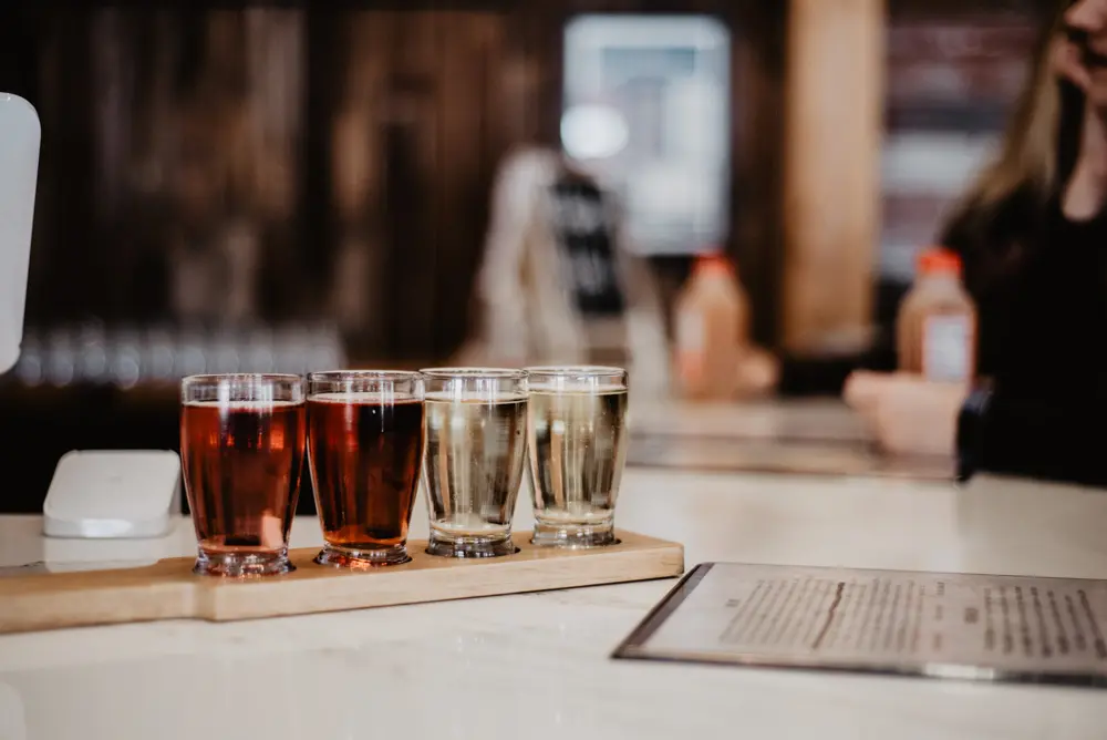 This image features a flight of cider or beer served in small glasses on a wooden tray, set on a polished bar counter. The drinks range in color from deep amber to light gold, suggesting a variety of flavors. The blurred background of a cozy establishment adds to the inviting and relaxed atmosphere of this tasting experience.