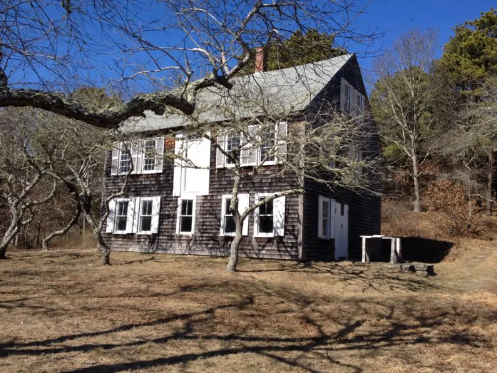 This image depicts a rustic wooden house with weathered shingles and white-trimmed windows, surrounded by leafless trees under a bright blue sky. The house sits on a dry, grassy patch of land with a backdrop of sparse woods, giving it a quiet, historic charm. The setting feels serene and rooted in nature, ideal for a peaceful retreat.