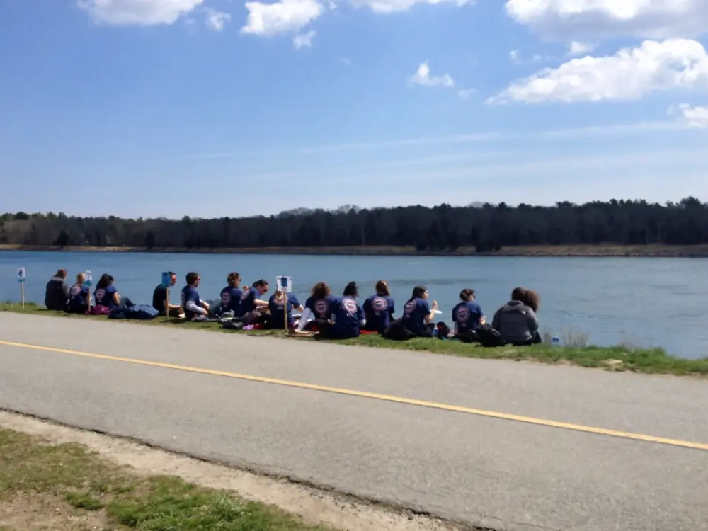 This image captures a group of people sitting on a grassy bank beside a calm canal, enjoying lunch under a partly cloudy sky. The group appears to be wearing matching shirts, suggesting a shared activity or event. The paved path alongside the canal adds a sense of accessibility to the peaceful outdoor setting.