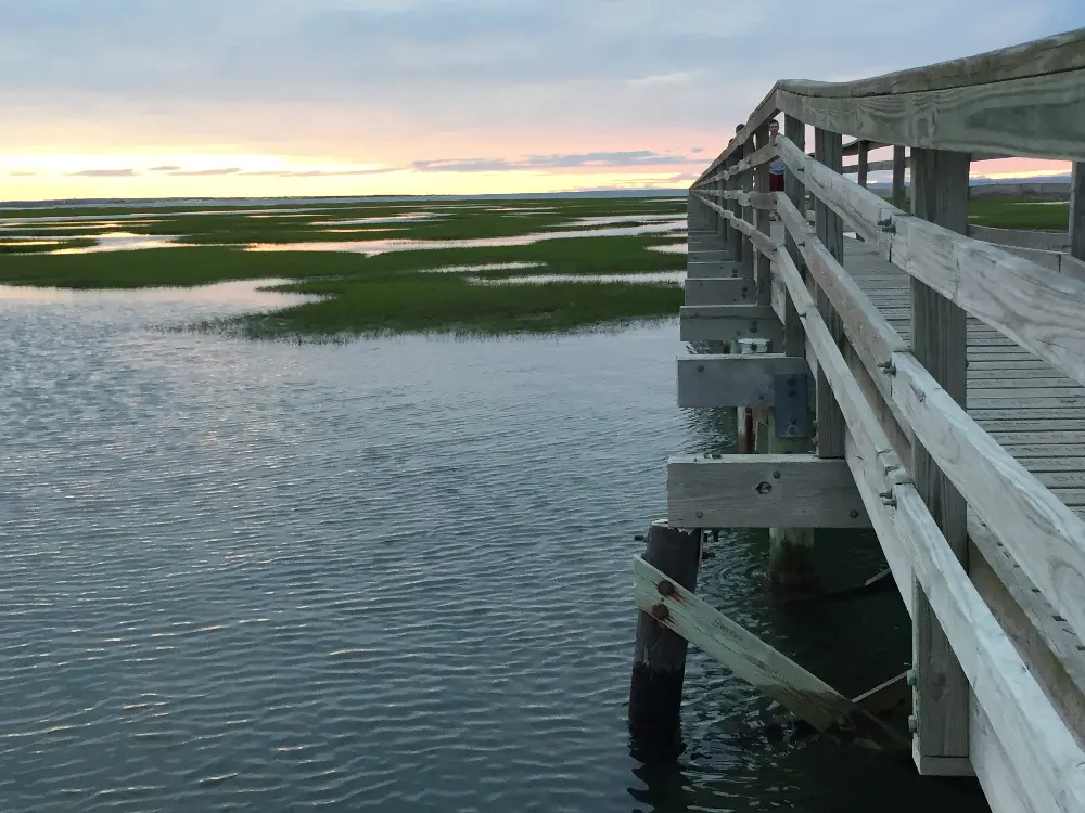 A wooden boardwalk extends over a calm body of water, leading toward a marshy area under a colorful sunset sky. The scene features reflections on the water and patches of green marshland, creating a serene and picturesque landscape.